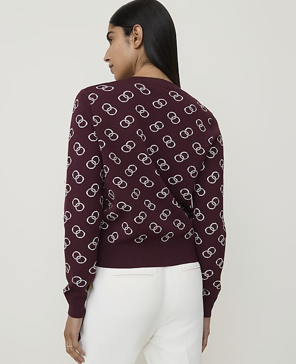 A woman in a maroon sweater with a white interlocking ring pattern stands against a plain background.