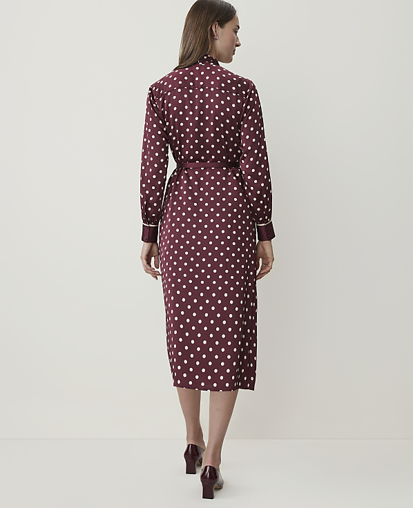A woman in a burgundy polka dot dress stands against a plain white background.