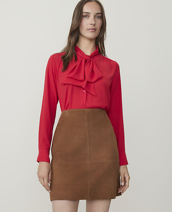 A woman in a red bow-neck blouse and brown suede skirt stands against a plain white background.