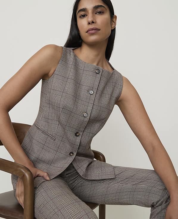 A woman in a plaid vest and pants sits on a wooden chair against a plain background.