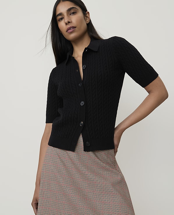 A woman in a black cable-knit polo shirt and a plaid skirt stands against a plain white background.