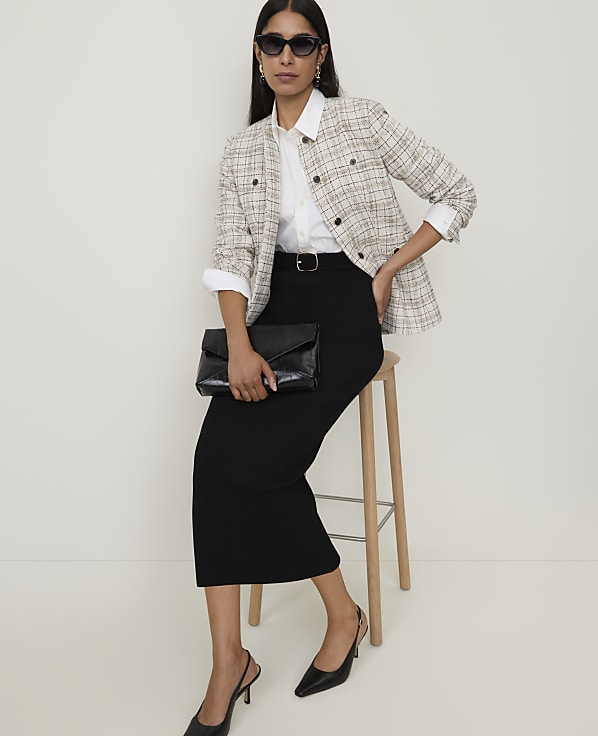 A woman in a tweed jacket and black skirt sits on a stool in front of a plain white background.