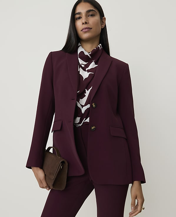 A woman in a burgundy blazer and matching pants holds a brown suede clutch in front of a plain white background.