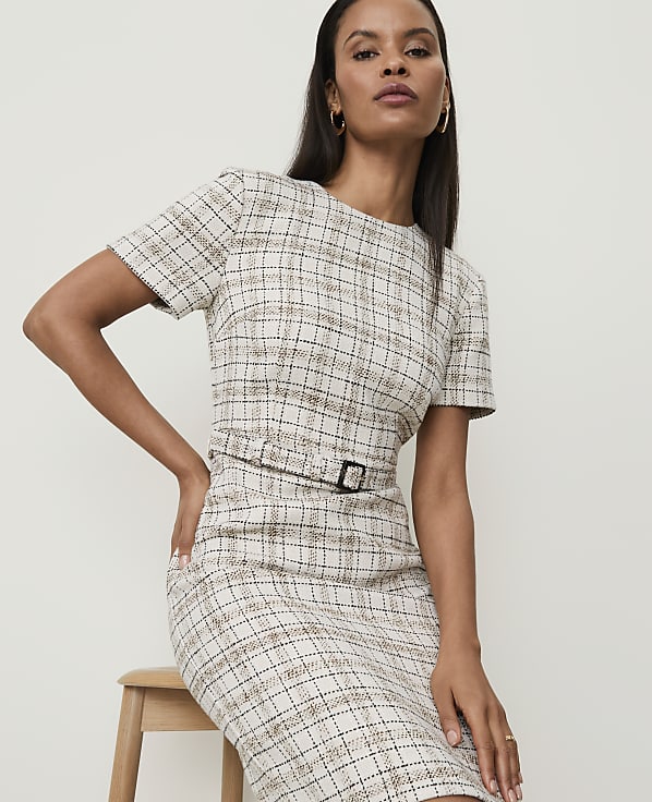 A woman in a tweed dress with a belt sits on a stool against a plain background.