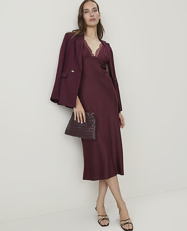 A woman in a burgundy slip dress and blazer holds a woven clutch in front of a plain, light-colored wall.