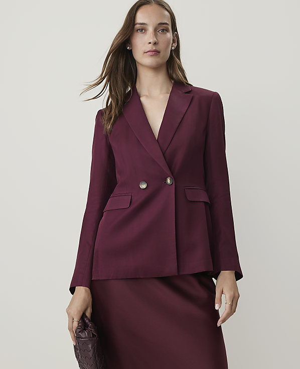 A woman in a burgundy blazer and skirt holds a textured clutch in front of a plain, light-colored background.