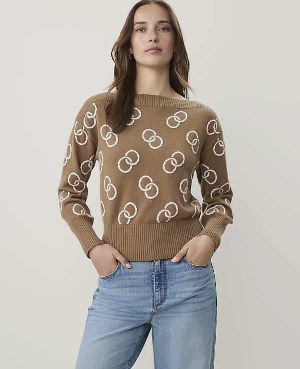 A woman in a tan sweater with a white interlocking ring pattern stands against a plain white background.
