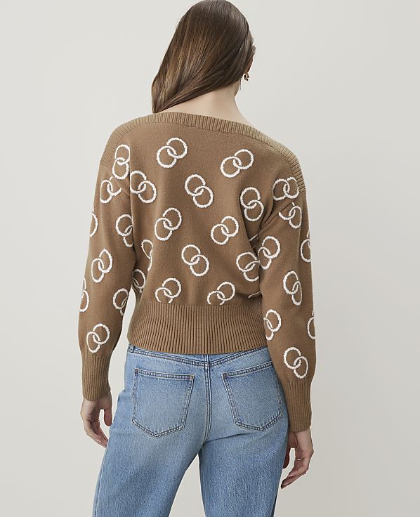 A woman in a brown sweater with a white interlocking ring pattern and blue jeans stands against a plain, light background.