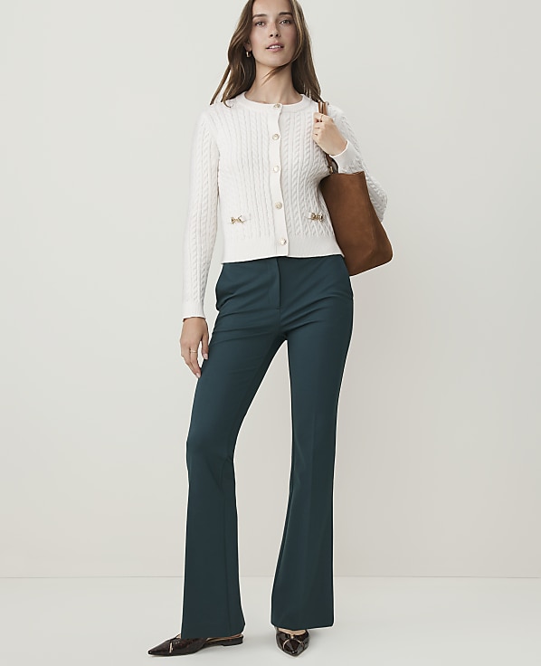 A woman in a white cardigan and teal flared pants stands against a plain, light background, carrying a brown tote bag.