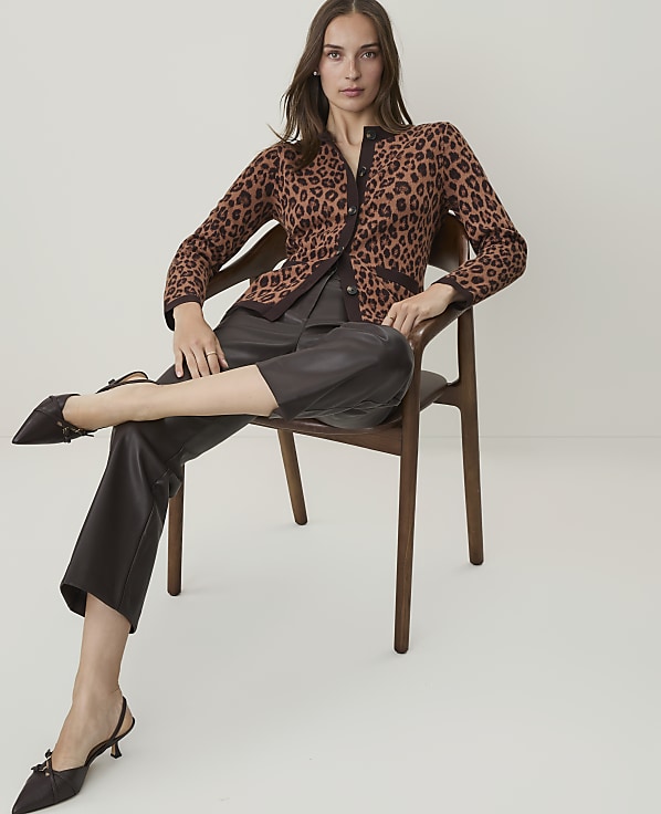A woman in a leopard print cardigan and dark pants sits on a wooden chair against a plain white background.