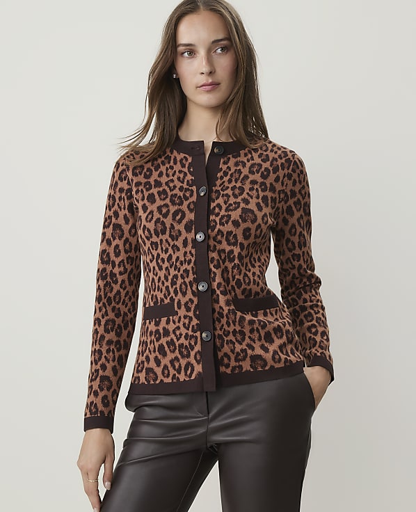 A woman in a leopard print cardigan and dark pants stands against a plain, light brown background.