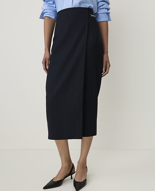 A person stands in a studio setting, wearing a pinstripe wrap skirt and a blue button-down shirt, with black slingback heels on their feet.