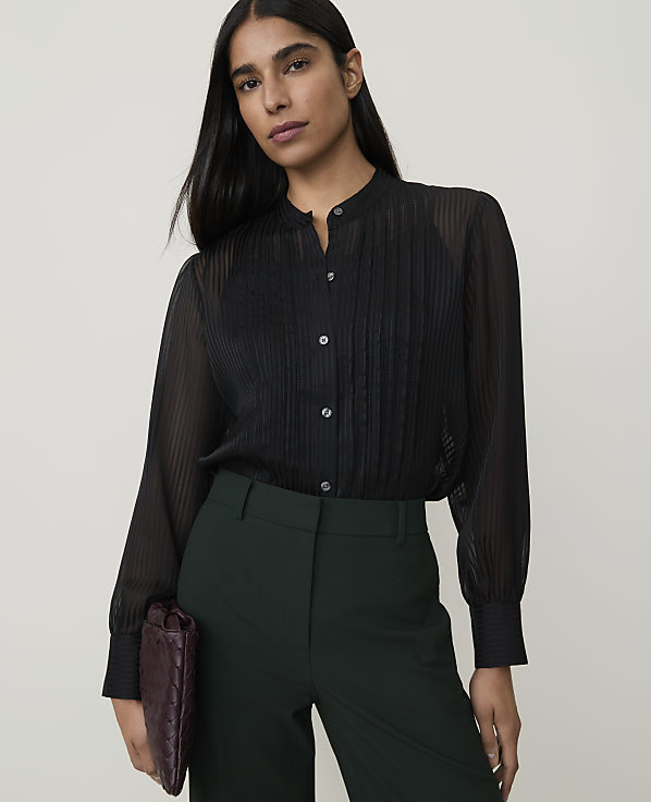 A woman in a sheer black blouse and dark green pants holds a burgundy clutch against a plain white background.