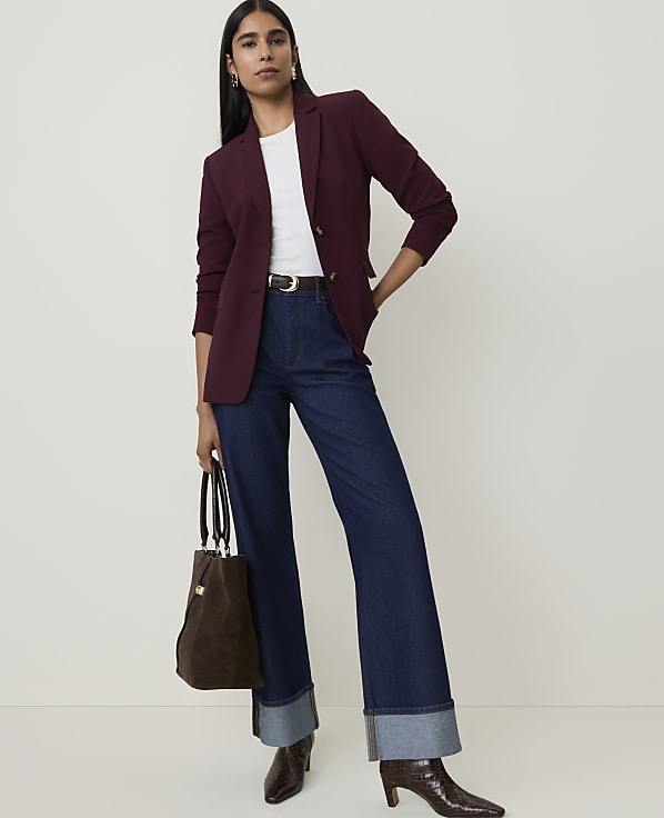 A woman in a burgundy blazer and dark wash wide-leg jeans stands against a plain white background, holding a brown tote bag.