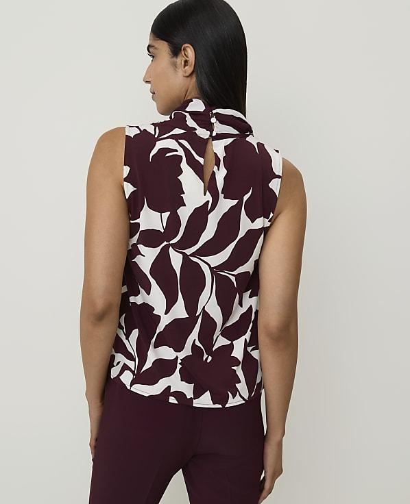 A woman in a floral sleeveless top and maroon pants stands against a plain white background.