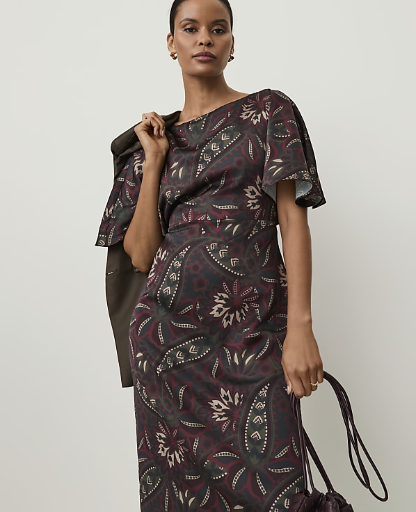 A woman in a patterned dress and jacket stands against a plain background, holding a handbag.