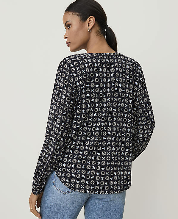 A woman in a patterned blouse and jeans stands against a plain background.