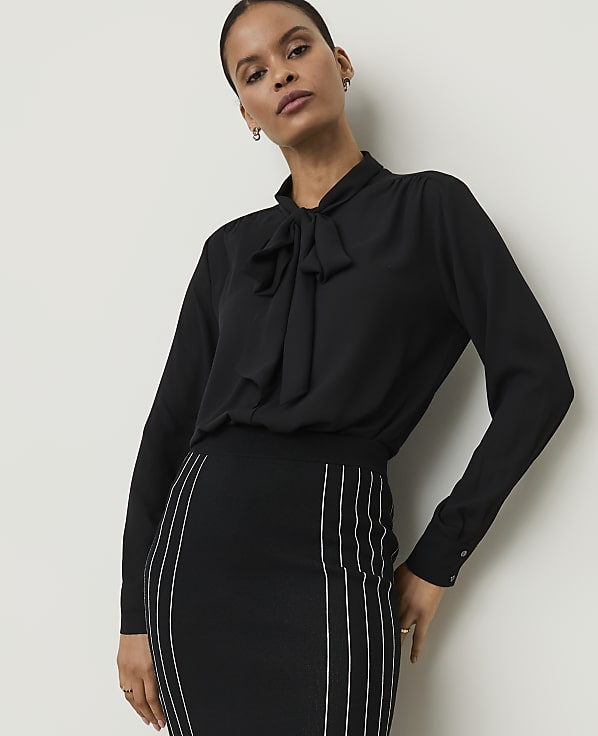 A woman in a black pussy-bow blouse and a striped pencil skirt stands against a plain white background.