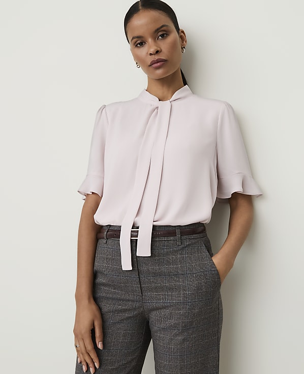 A woman in a light pink bow-neck blouse and plaid trousers stands against a plain white background.