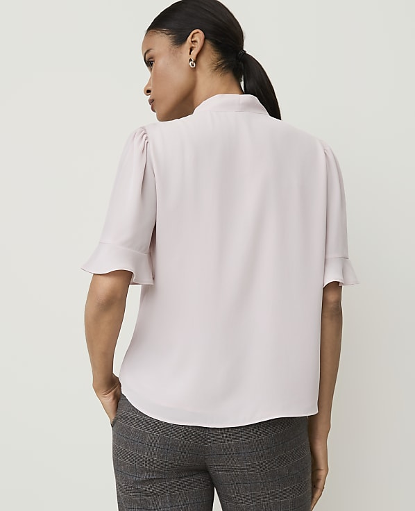A woman in a light purple blouse and plaid pants stands with her back to the camera against a plain white background.