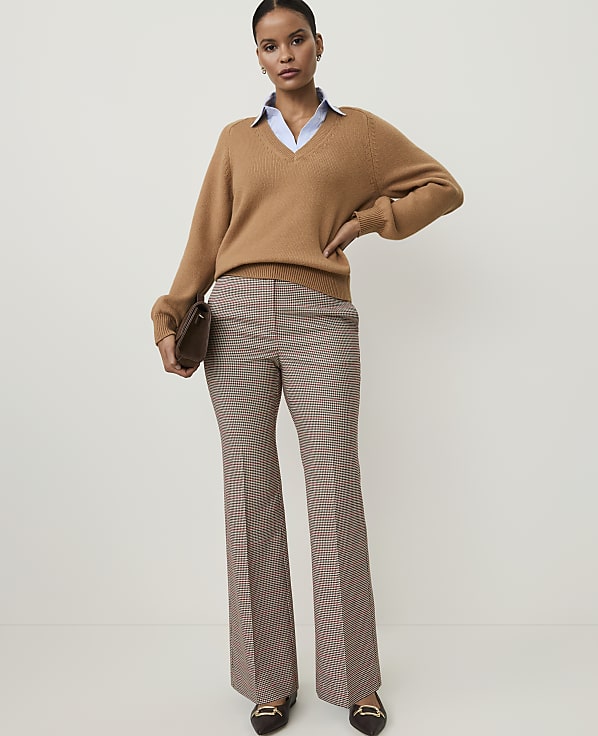A woman in a camel v-neck sweater, light blue collared shirt, and plaid flared pants stands against a plain, light background, holding a brown clutch.
