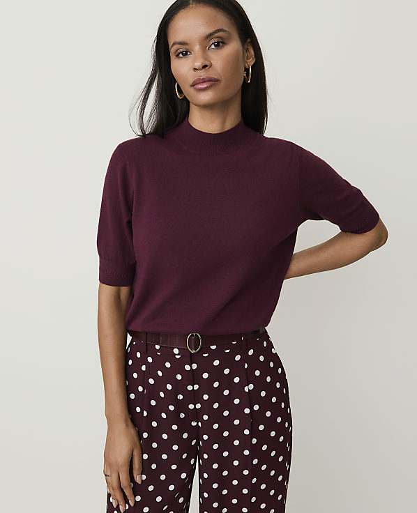 A woman in a burgundy mock-neck sweater and polka-dot pants stands against a plain, light background.