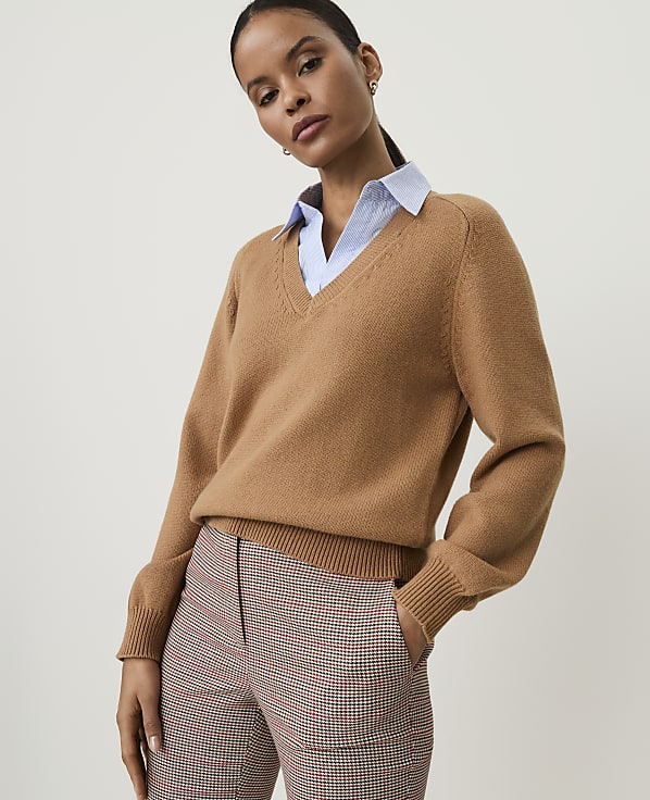 A woman in a camel v-neck sweater over a light blue collared shirt and plaid pants stands against a plain, light background.