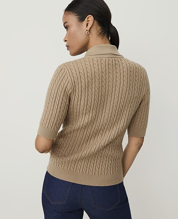 A woman in a tan cable-knit sweater and dark jeans stands against a plain beige background.