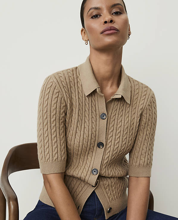 A woman in a cable-knit cardigan sits in front of a plain, light-colored background.
