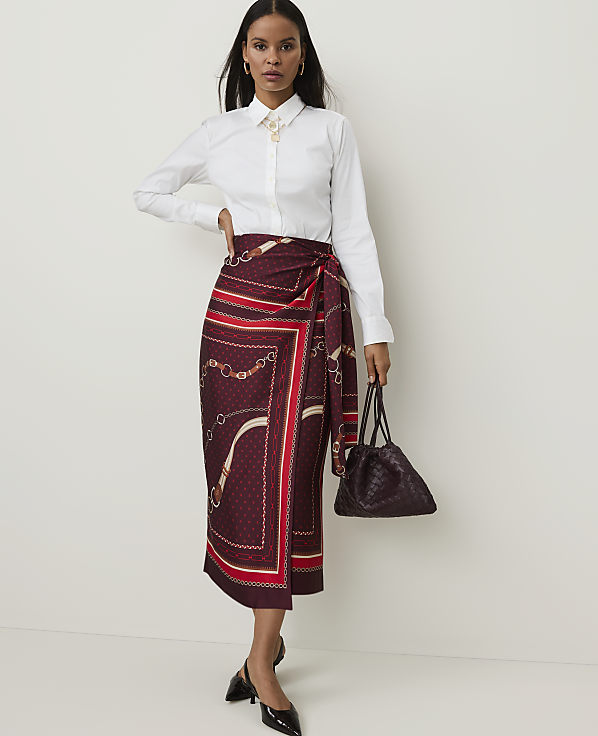 A woman in a white collared shirt and a patterned wrap skirt stands against a plain white background, holding a dark handbag.