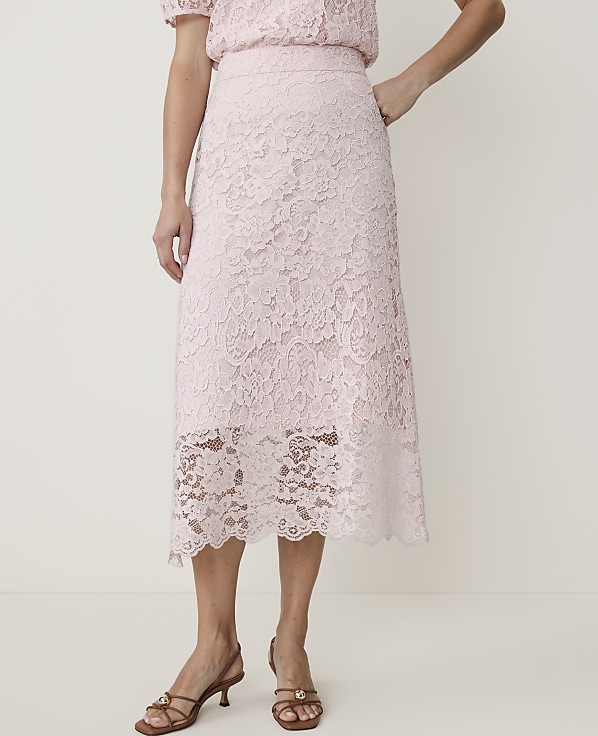 A woman in a light pink lace skirt and matching top stands against a plain white background, her feet adorned with brown heeled sandals.