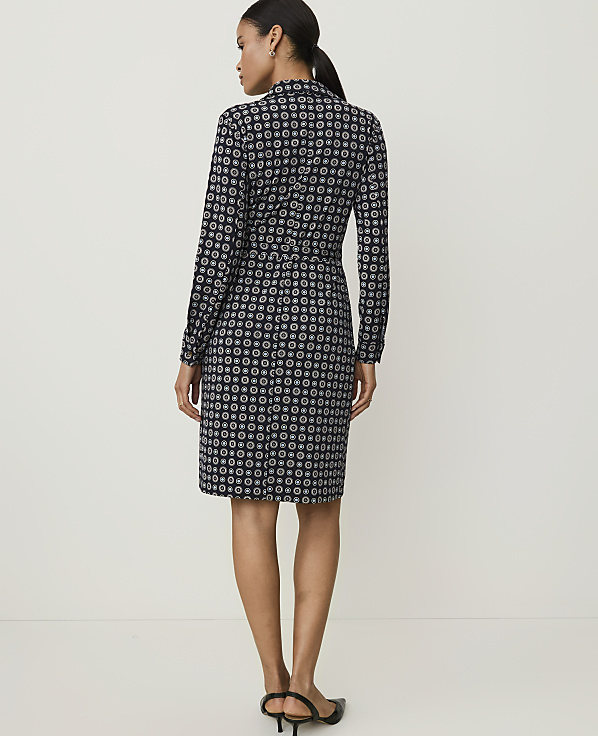 A woman in a patterned dress stands against a plain, light-colored background.