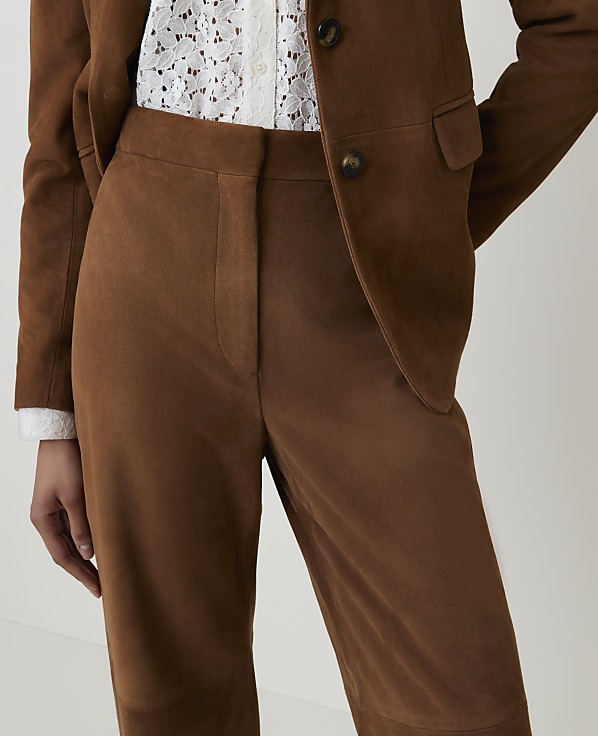 A person wears a brown suede suit with a white lace top, against a plain background.