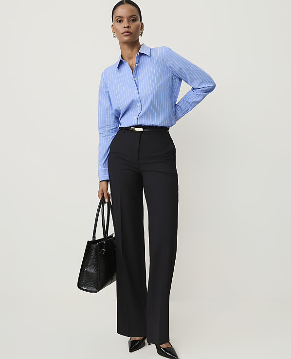 A woman in a blue striped shirt and black pants holds a black handbag against a plain white background.