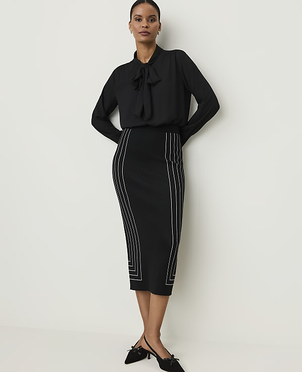 A woman in a black bow-neck blouse and a black pencil skirt with white geometric detailing stands against a plain white background.