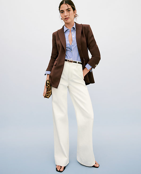 A woman in a brown blazer, striped shirt, and white wide-leg pants stands against a plain blue background, holding a leopard-print clutch.