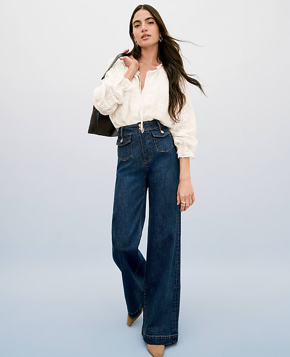 A woman in a white blouse and wide-leg jeans stands against a light blue background, with a dark jacket draped over her shoulder.