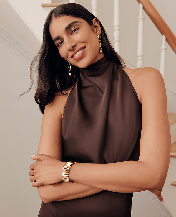 A smiling woman in a brown halter dress and jewelry poses in front of a wooden staircase.