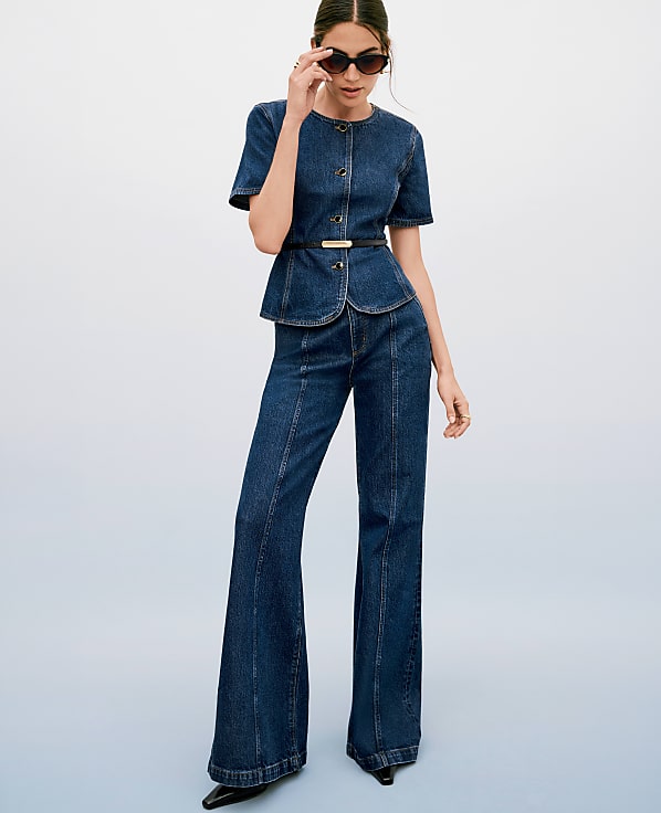 A woman in a denim peplum top and flared jeans poses against a gradient blue background.