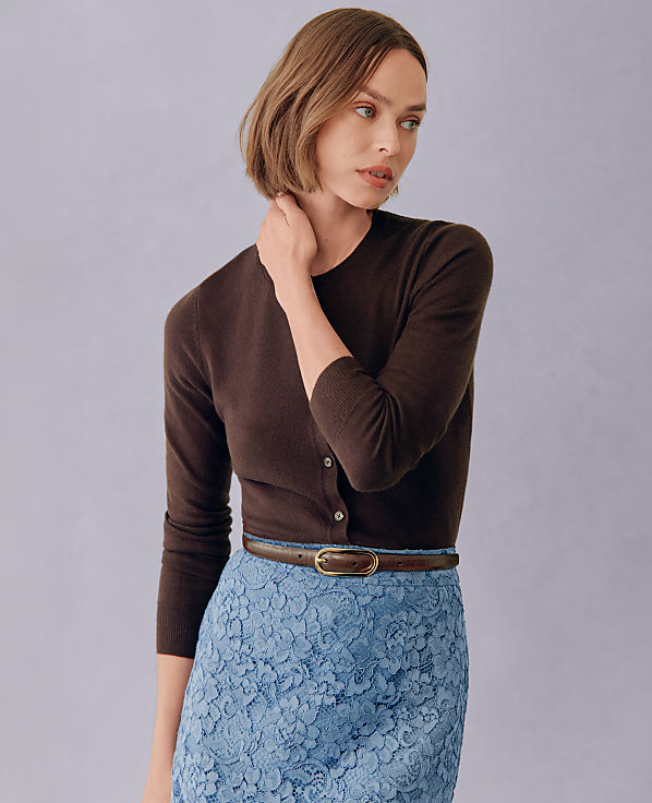 A woman in a brown cardigan and blue lace skirt stands against a plain purple background.