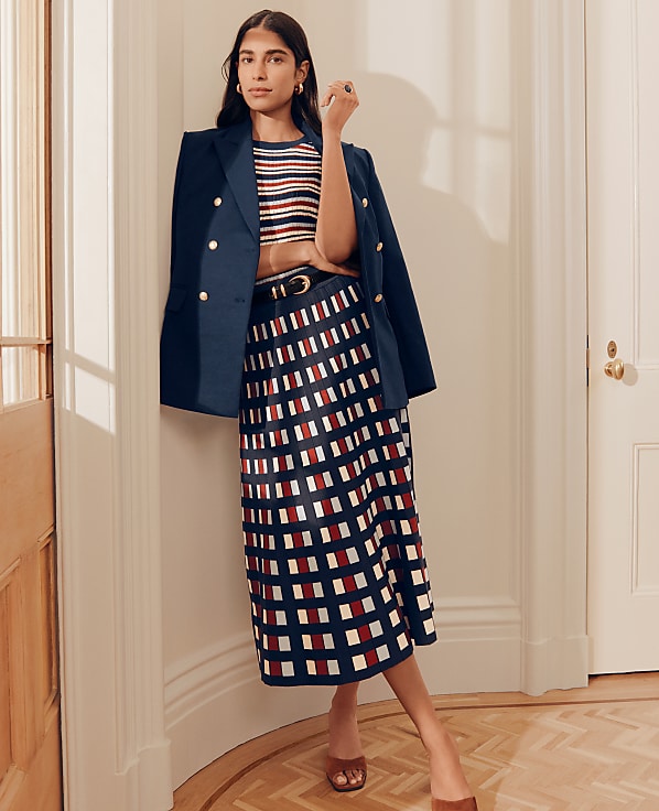 A woman in a striped top, navy blazer, and patterned skirt stands in a hallway with a wooden floor and white doors.
