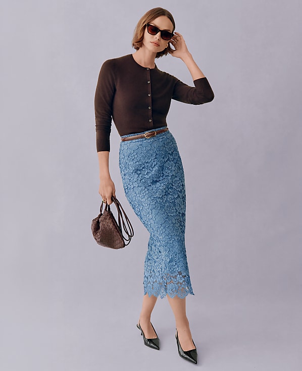 A woman in a brown cardigan and blue lace skirt poses with a handbag against a plain background.
