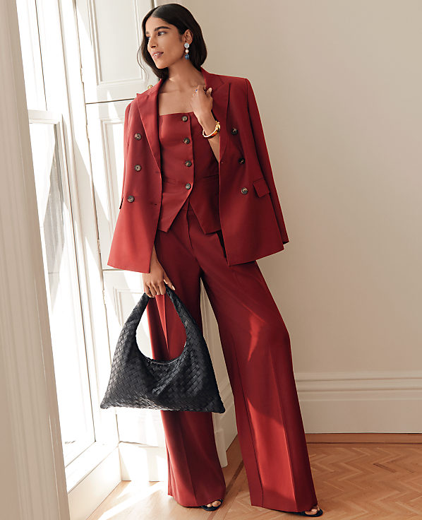 A woman in a red pantsuit and a black woven bag stands by a window with white blinds, against a plain wall.