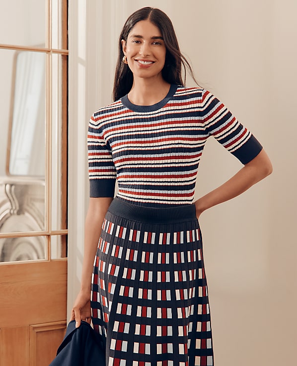 A smiling woman in a striped top and patterned skirt stands in a well-lit room with a doorway to her left.