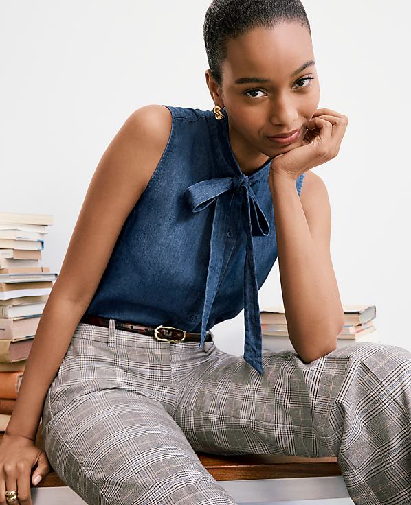 A woman in a denim bow-neck top and plaid pants rests her chin on her hand, with a stack of books behind her.