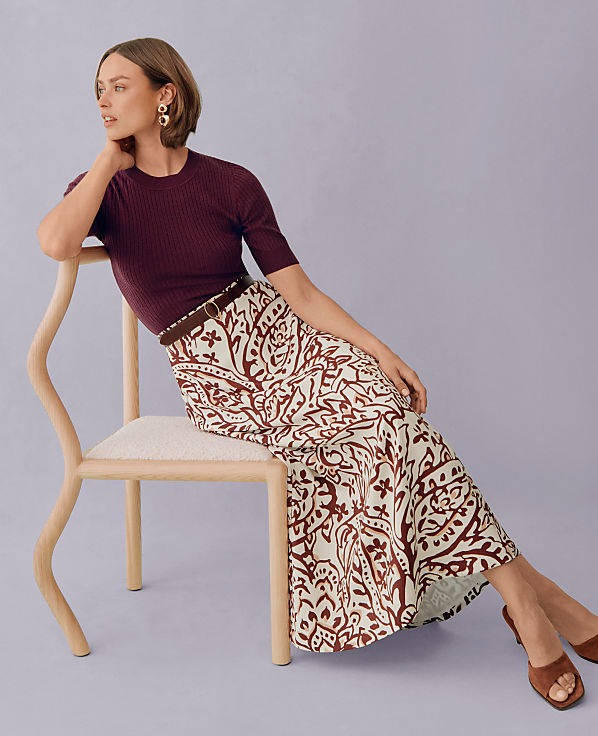 A woman in a patterned skirt and maroon top sits on a chair against a lavender background.