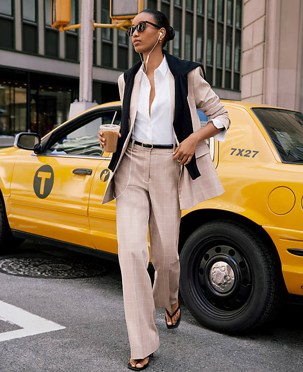 A woman in a tan plaid suit walks past a yellow taxi in a city street.