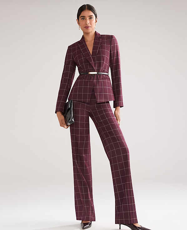 A woman in a burgundy plaid suit with a black clutch stands against a plain, light gray background.