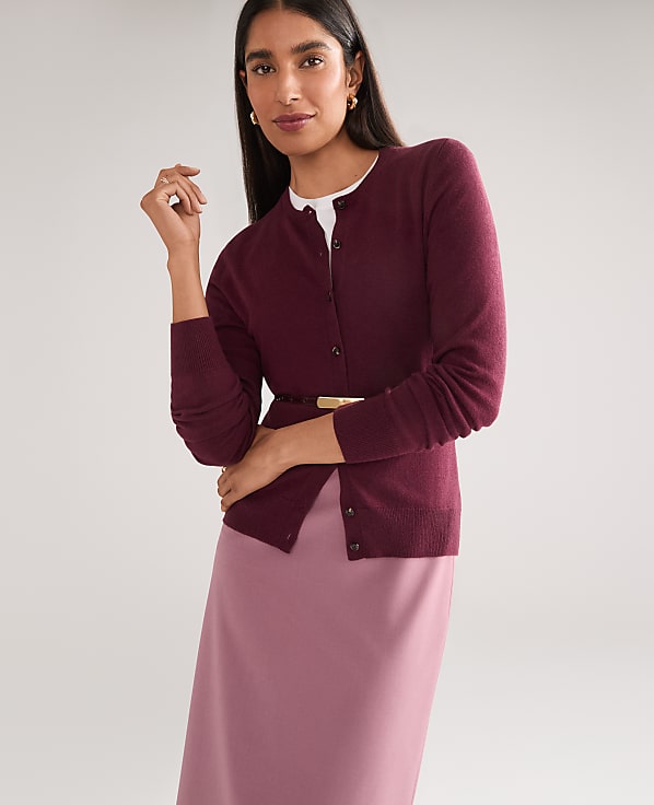 A woman in a burgundy cardigan and pink skirt stands against a plain gray background.