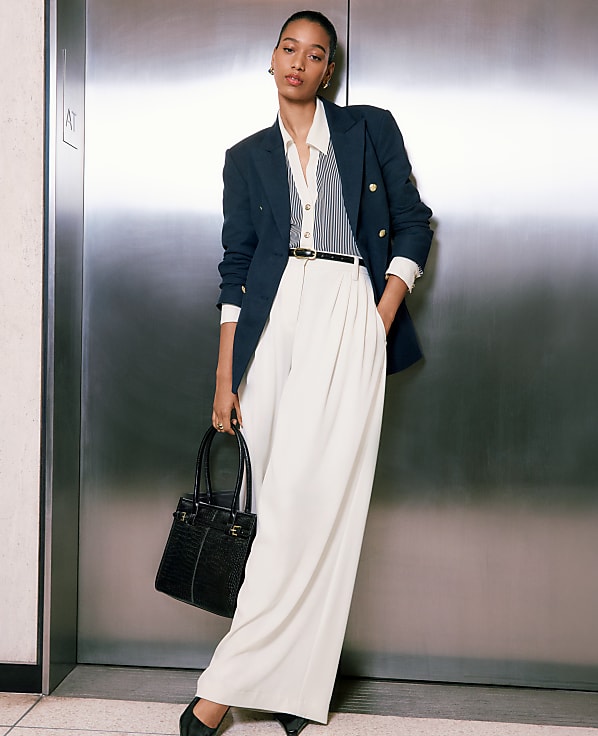 A woman in a navy blazer and white pants stands in front of a brushed metal elevator door, holding a black handbag.